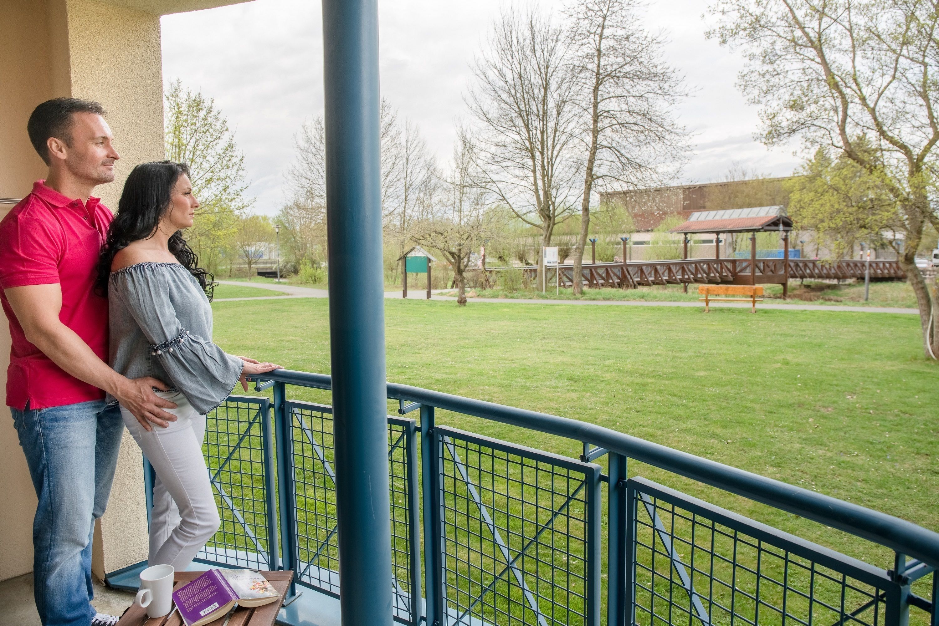 A couple standing on a balcony overlooking a grassy area with a playground and a building in the background.