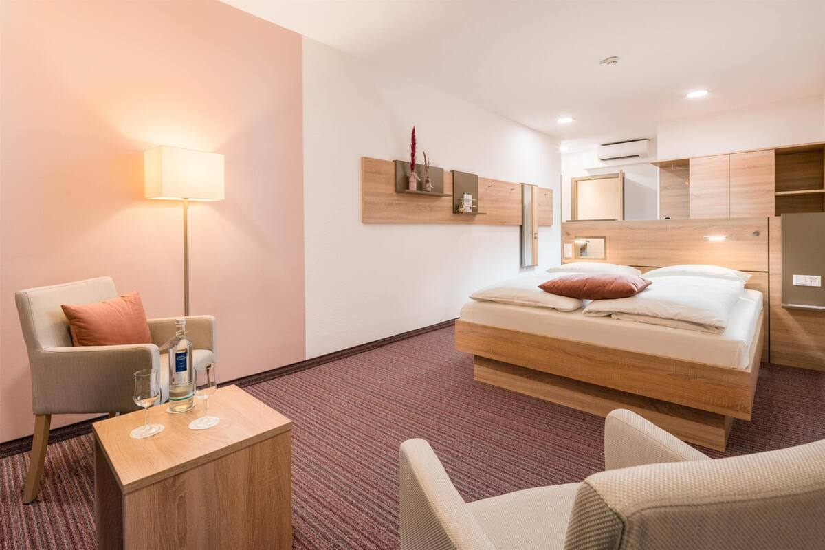 A hotel room with a bed, a chair, a table, a lamp, and a wall-mounted shelf with decorative items.