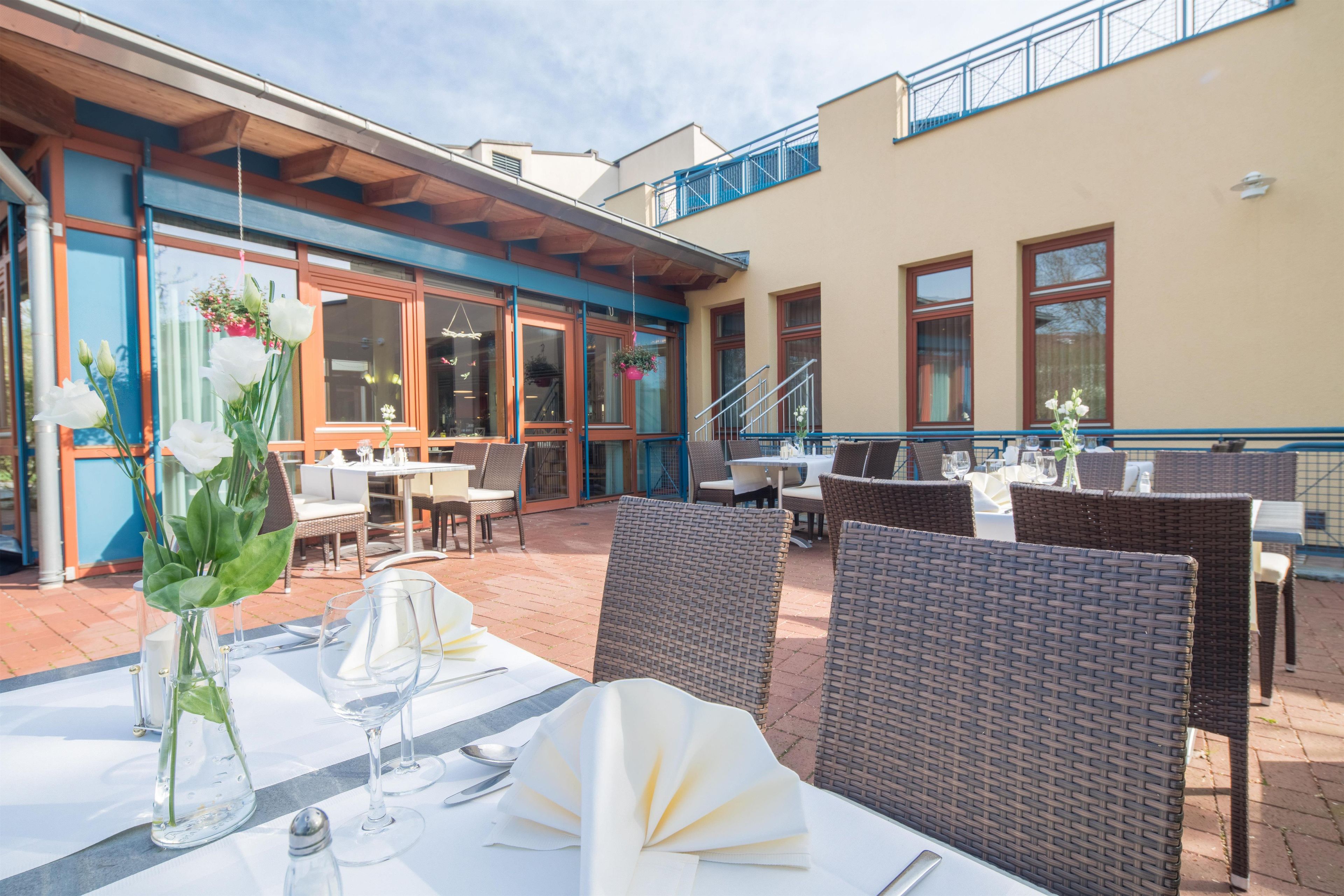 An outdoor dining area with wicker chairs and tables, a covered section with a wooden ceiling, and a building with large windows and a blue railing.