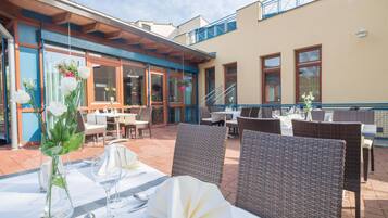 An outdoor dining area with wicker chairs and tables, a covered section with a wooden ceiling, and a building with large windows and a blue railing.