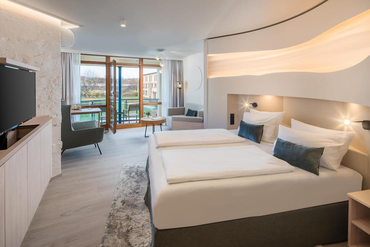 A modern hotel room with two beds, a TV, a balcony with a table and chairs, and a view of buildings.