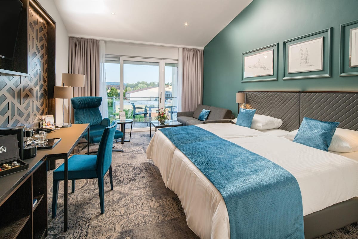 A hotel room with a large bed, a desk with a computer, a chair, a sofa, and a balcony with a view.