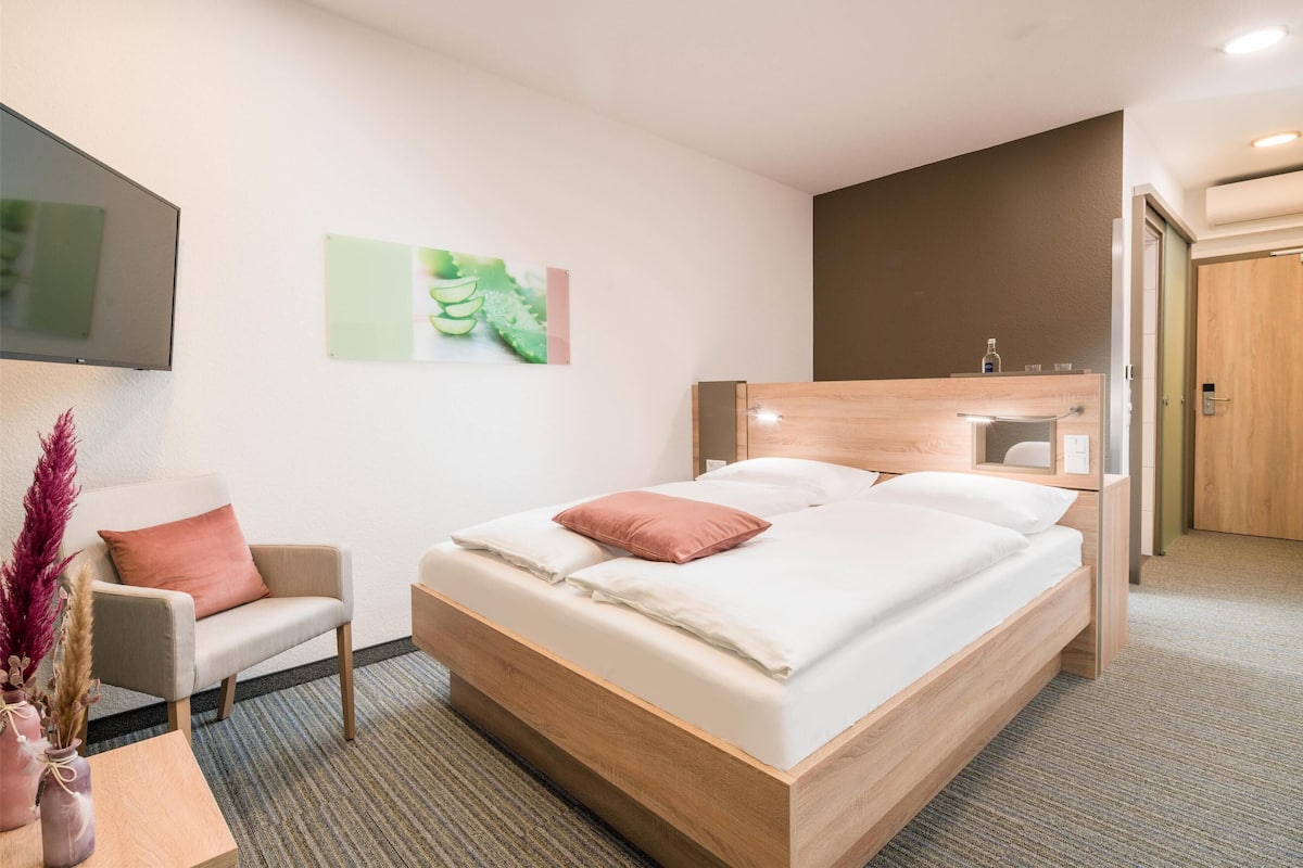 A modern hotel room with a wooden bed, a small desk, a chair, and a flat-screen TV mounted on the wall.