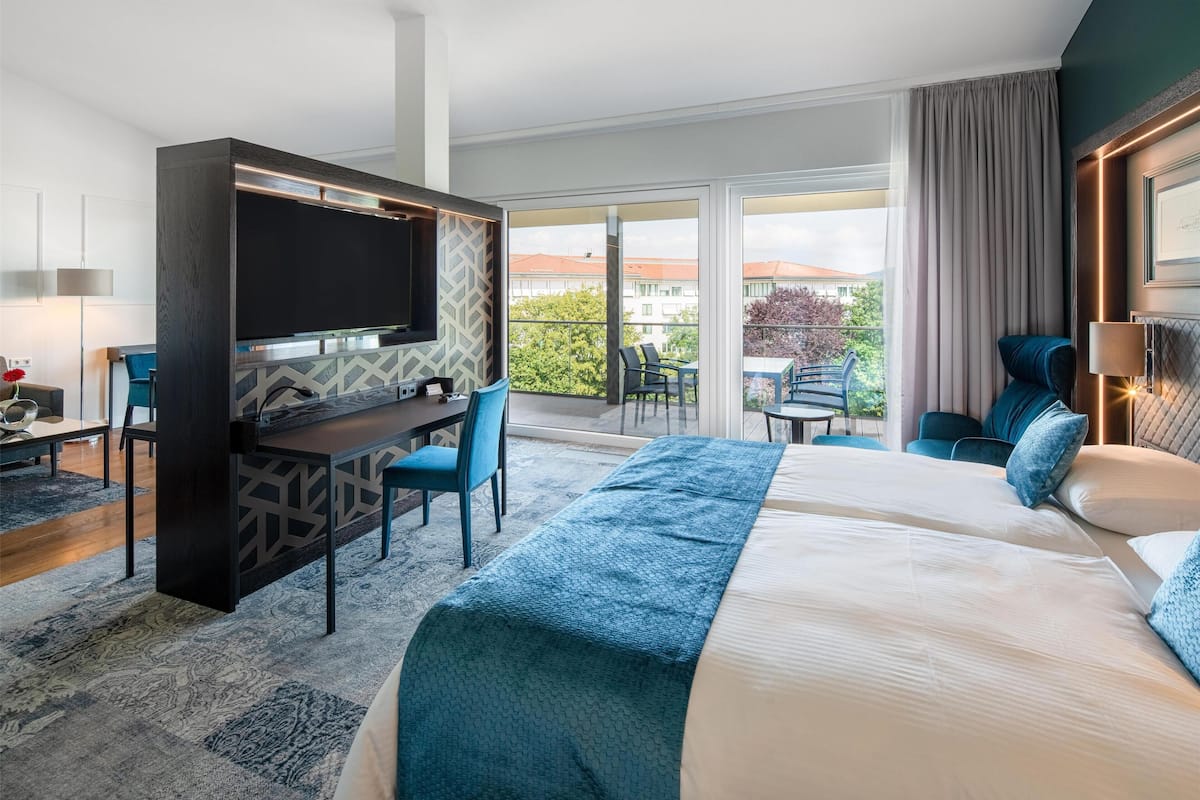 A hotel room with a large bed, a desk with a TV, a balcony with a table and chairs, and a view of buildings and trees.