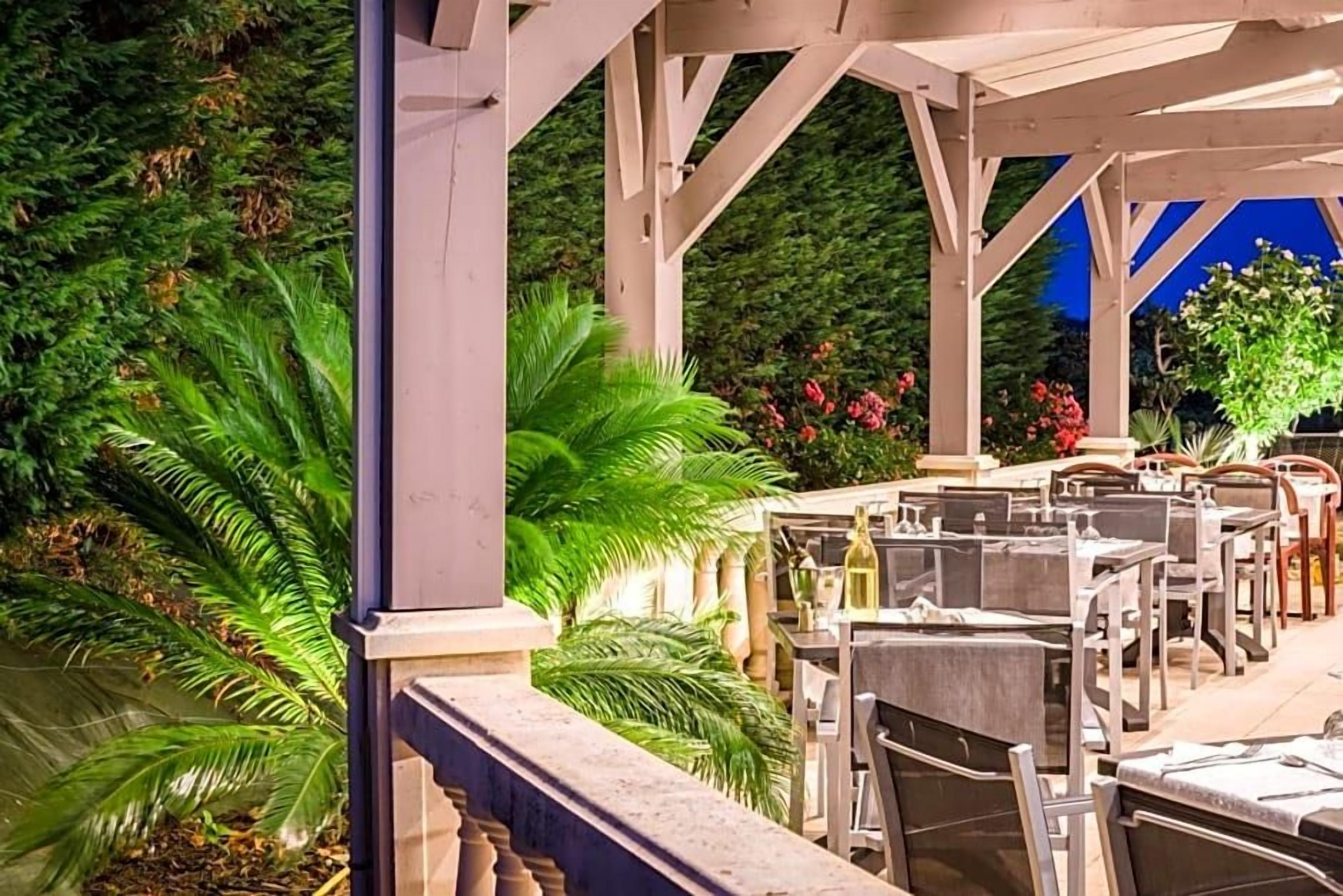 An outdoor patio with tables and chairs, surrounded by lush greenery and flowering plants.