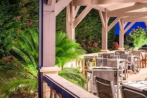 An outdoor patio with tables and chairs, surrounded by lush greenery and flowering plants.