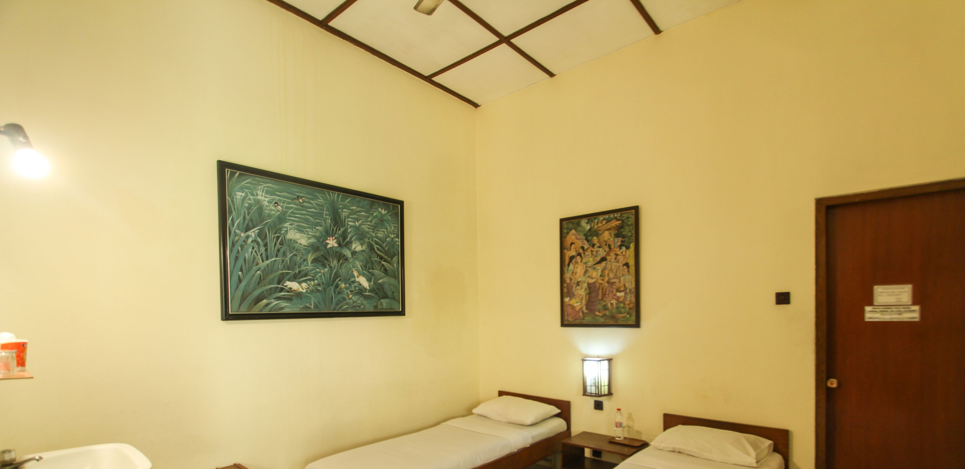 A small room with two single beds, a nightstand with a lamp, and two wall-mounted paintings.