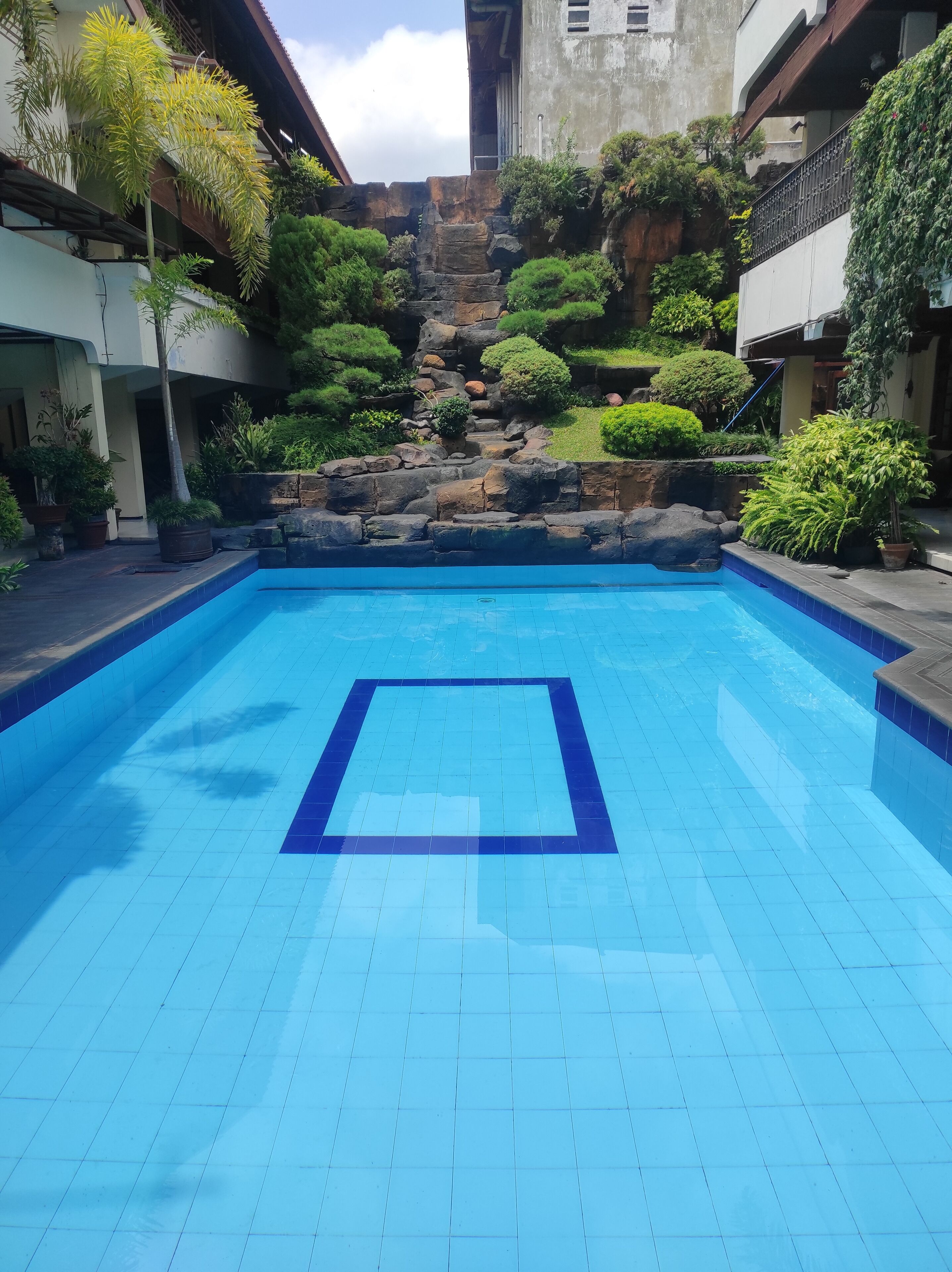 A swimming pool with a tiled deck, surrounded by lush greenery and a rocky backdrop.