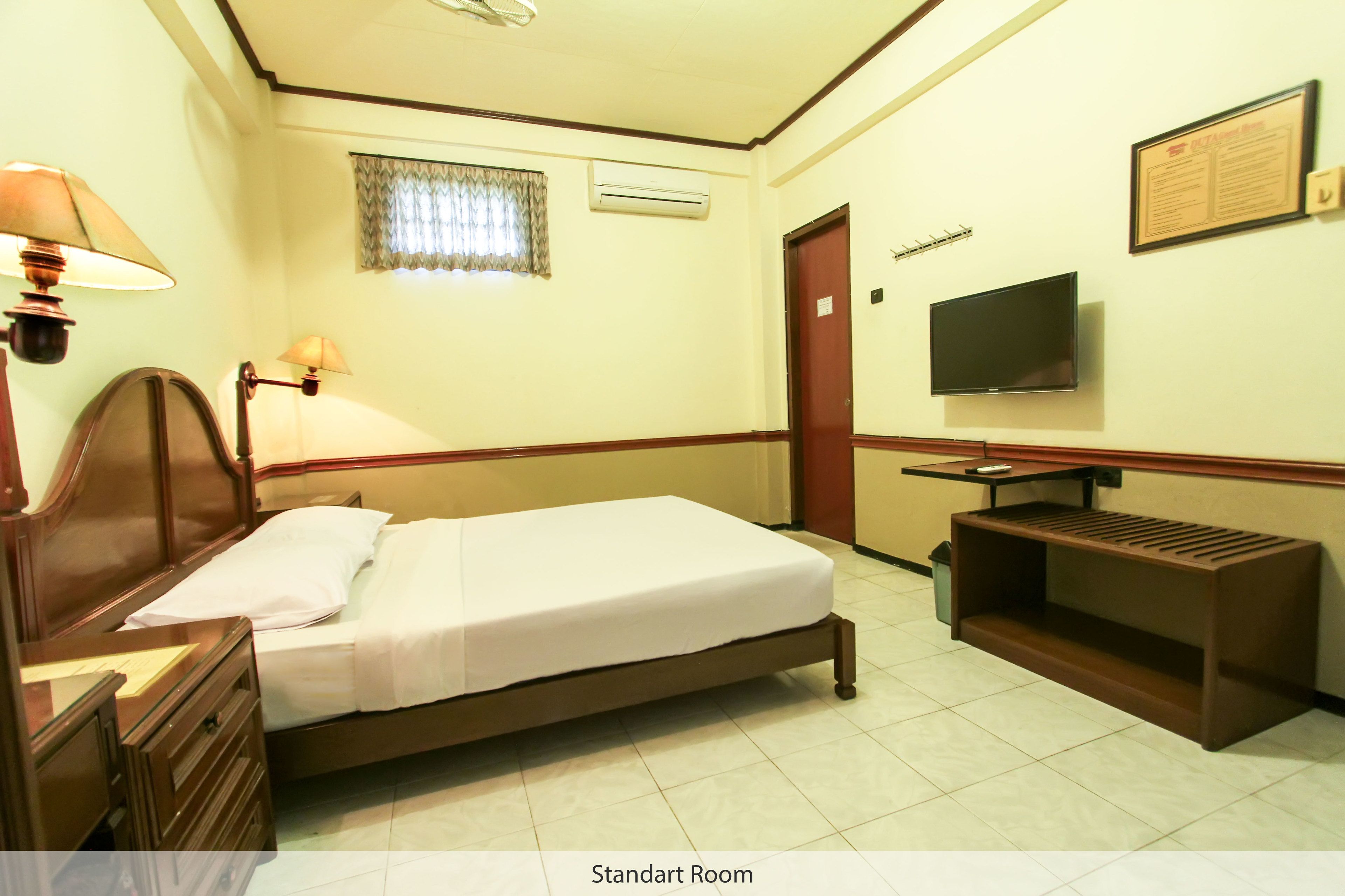 A standard room with a bed, bedside table, lamp, TV, and a small shelf.