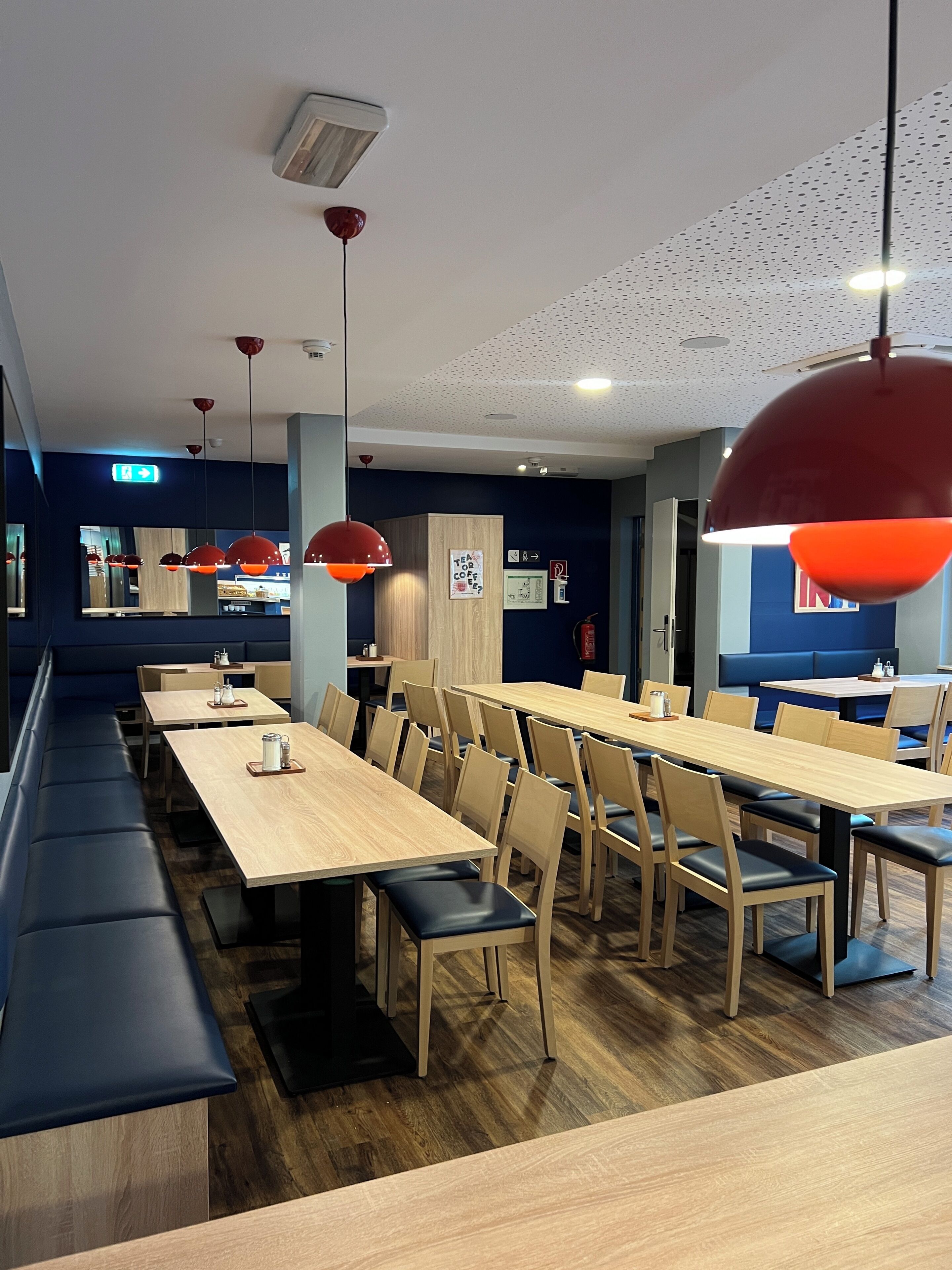A modern restaurant with long wooden tables, black cushioned benches, and red pendant lighting.