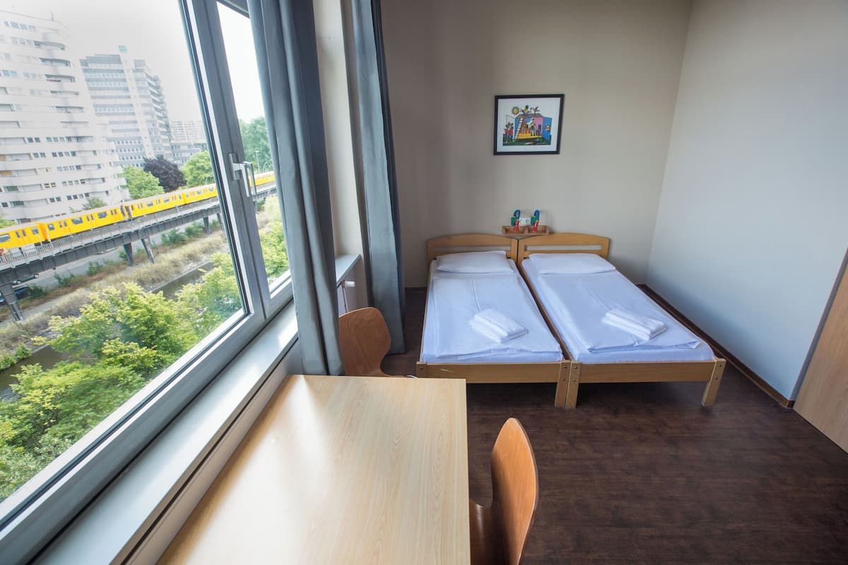Quarto de hotel com uma secretária de madeira, duas camas de solteiro, uma cadeira e janela com vista para um trem e vegetação.