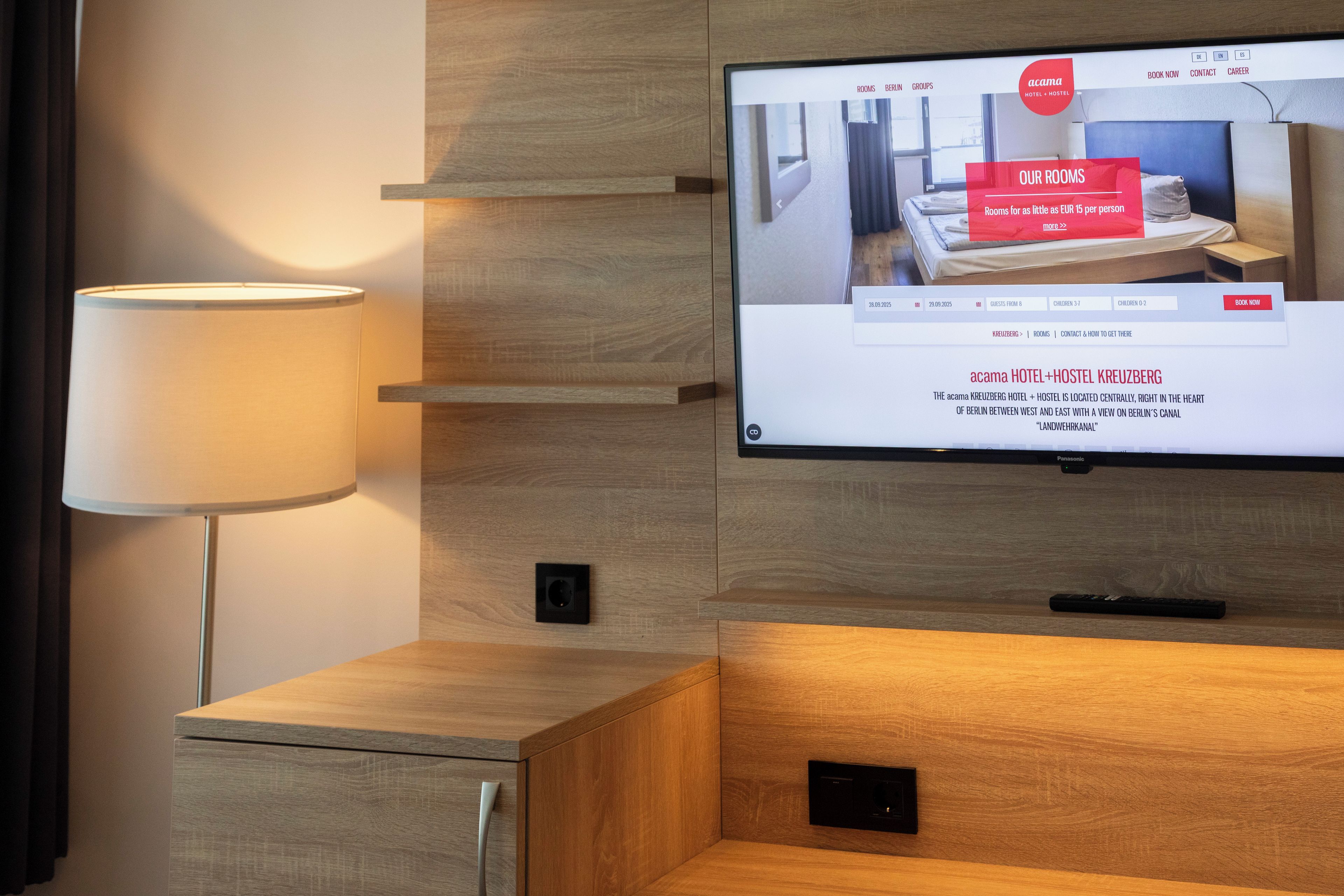 A hotel room with a television displaying a booking interface, a lamp, and a wooden cabinet.