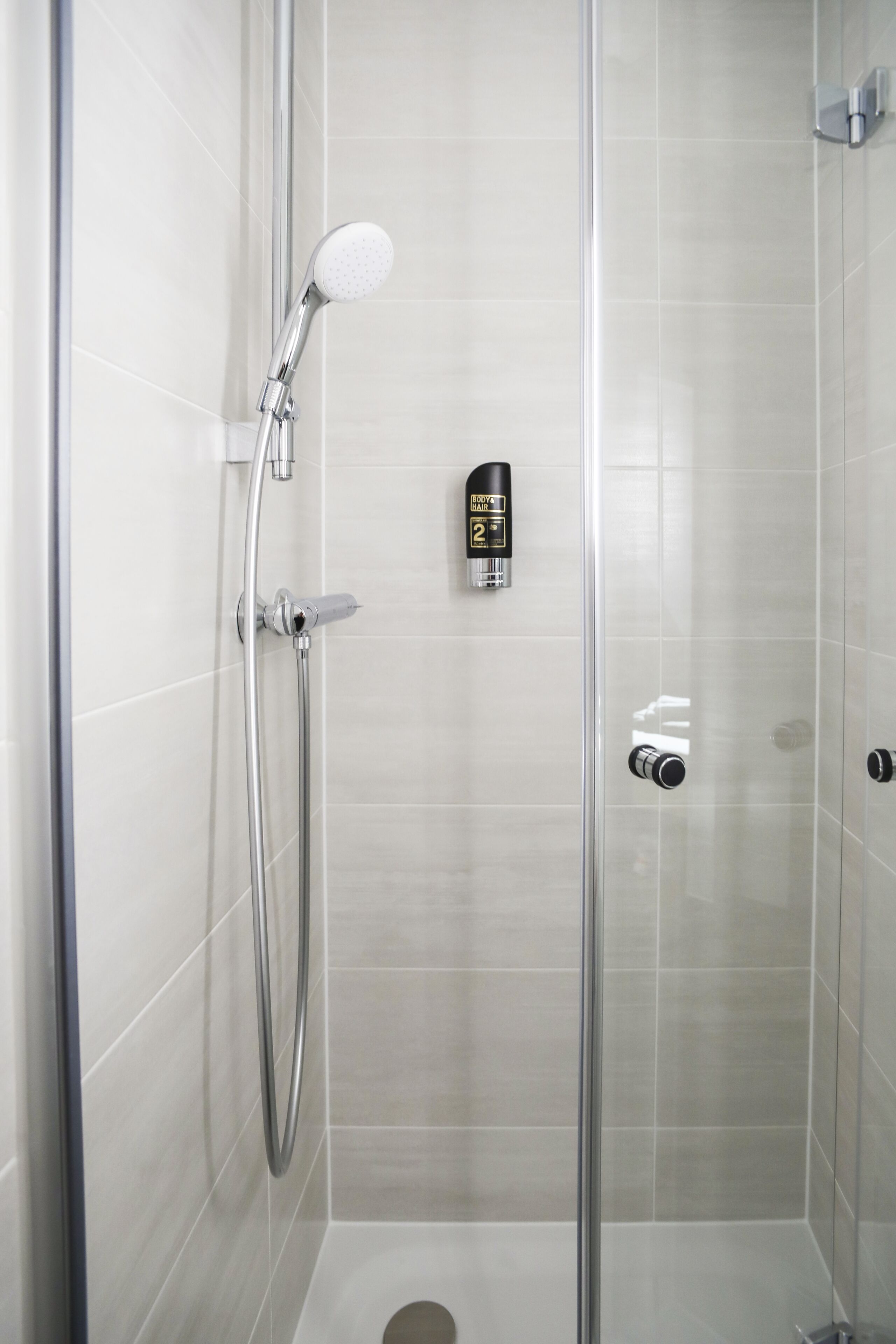 A glass-enclosed shower stall with a handheld showerhead, a bottle of shampoo, and a soap dispenser on the wall.