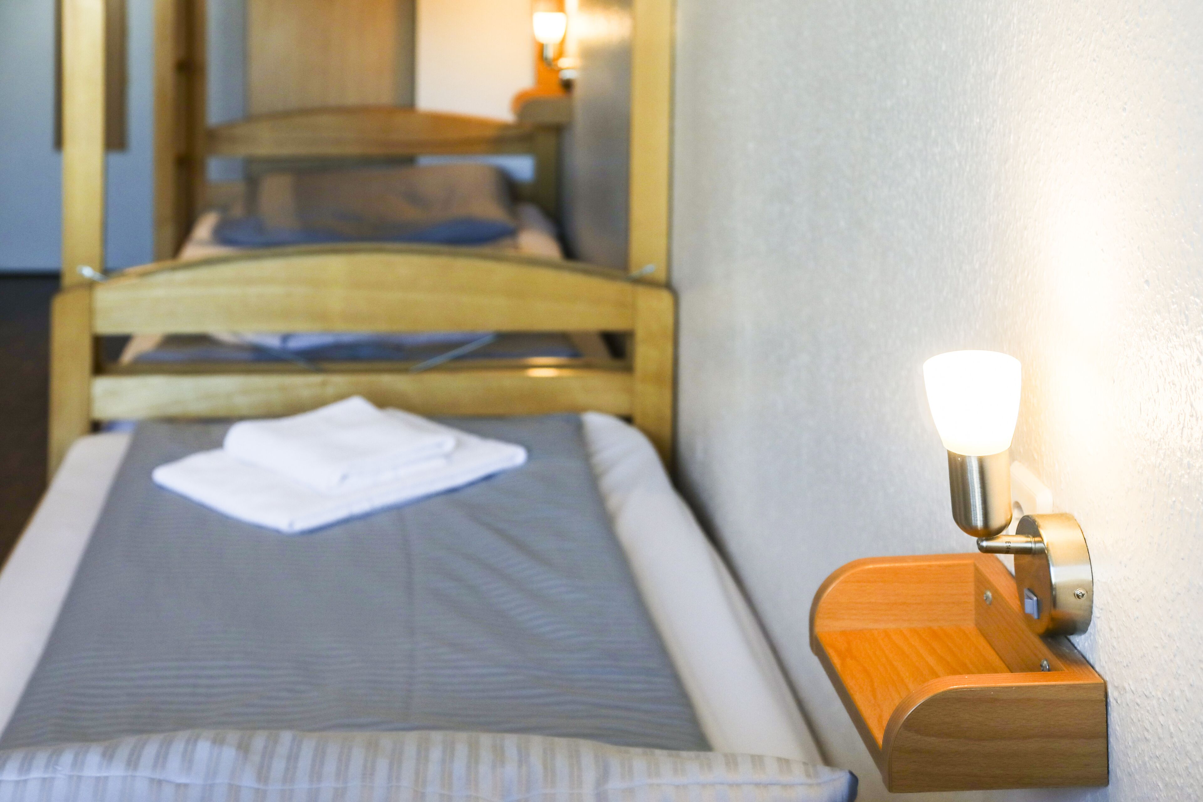 Un lit simple avec un matelas gris et des draps blancs, une tête de lit en bois et une table de chevet avec une lampe.