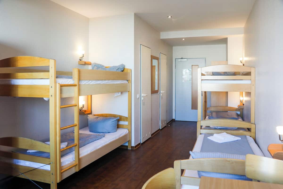 Quarto com beliches, uma escrivaninha e uma cadeira. Portas e um corredor são visíveis.
