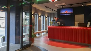 Un área de recepción moderna con un mostrador rojo, ventanas amplias y una pantalla de televisión mostrando una escena nocturna.