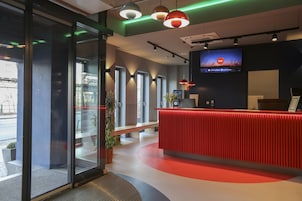 Un área de recepción moderna con un mostrador rojo, ventanas amplias y una pantalla de televisión mostrando una escena nocturna.