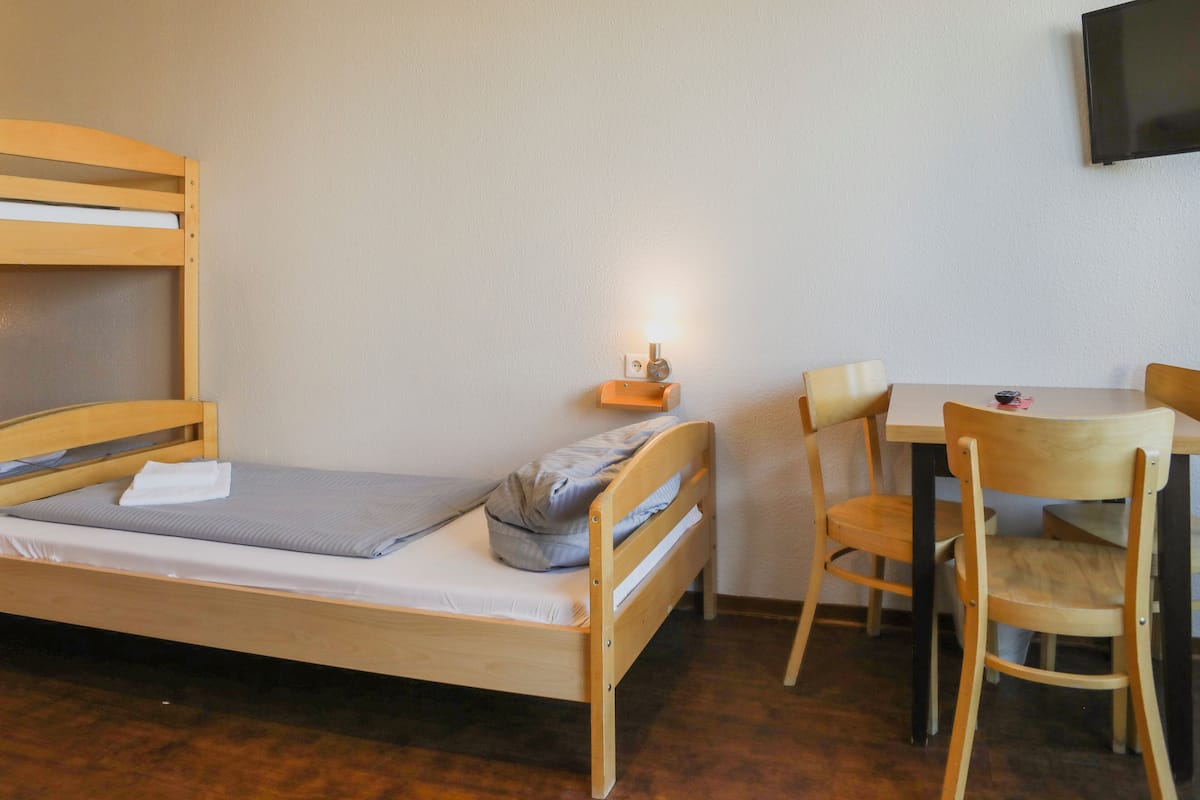 Uma cama de solteiro com cabeceira de madeira, mesa de cabeceira com abajur, uma cadeira e uma pequena mesa com outra cadeira.
