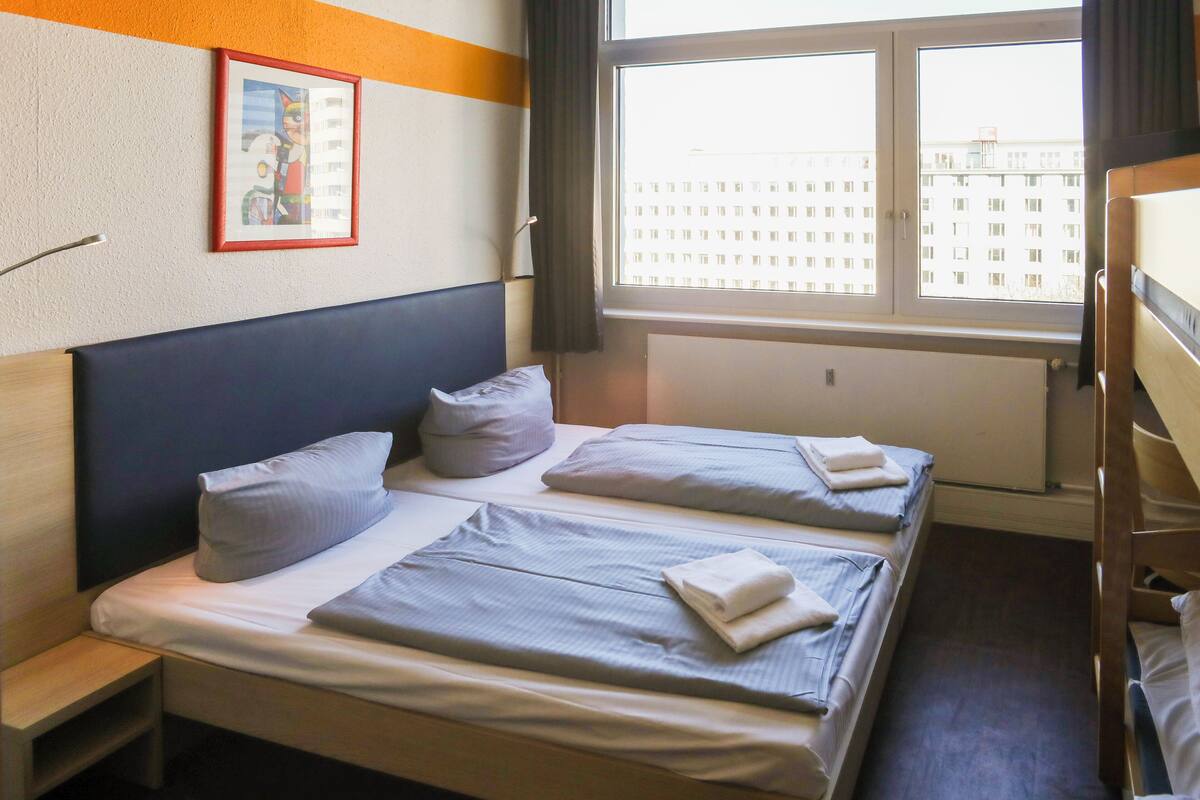 Quarto de hotel com duas camas, uma janela grande, um radiador e um quadro na parede.