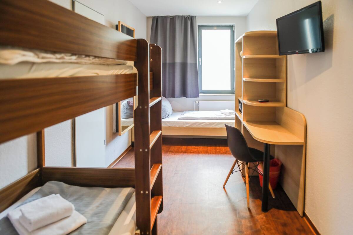 Quarto de hotel compacto com beliche, uma cama de solteiro, escrivaninha com cadeira, televisão fixada na parede e janela com cortinas.