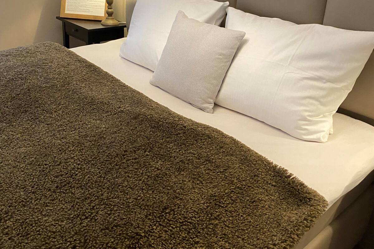 A neatly made bed with a grey blanket, white pillows, and a small bedside table with a lamp and a framed item.