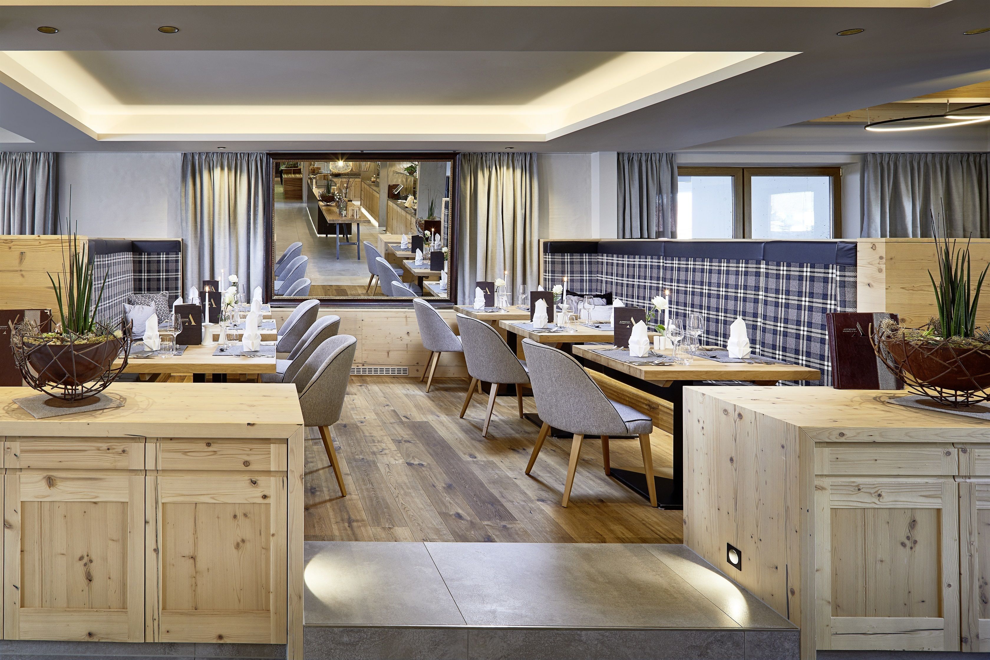 Un comedor moderno con mesas y sillas de madera, una escalera central y ventanas amplias con cortinas.