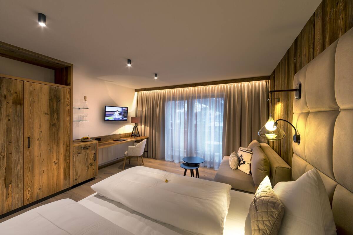 Une chambre d’hôtel moderne avec une armoire en bois, un bureau avec une chaise, un canapé, un lit avec une literie blanche et une grande fenêtre avec des rideaux.