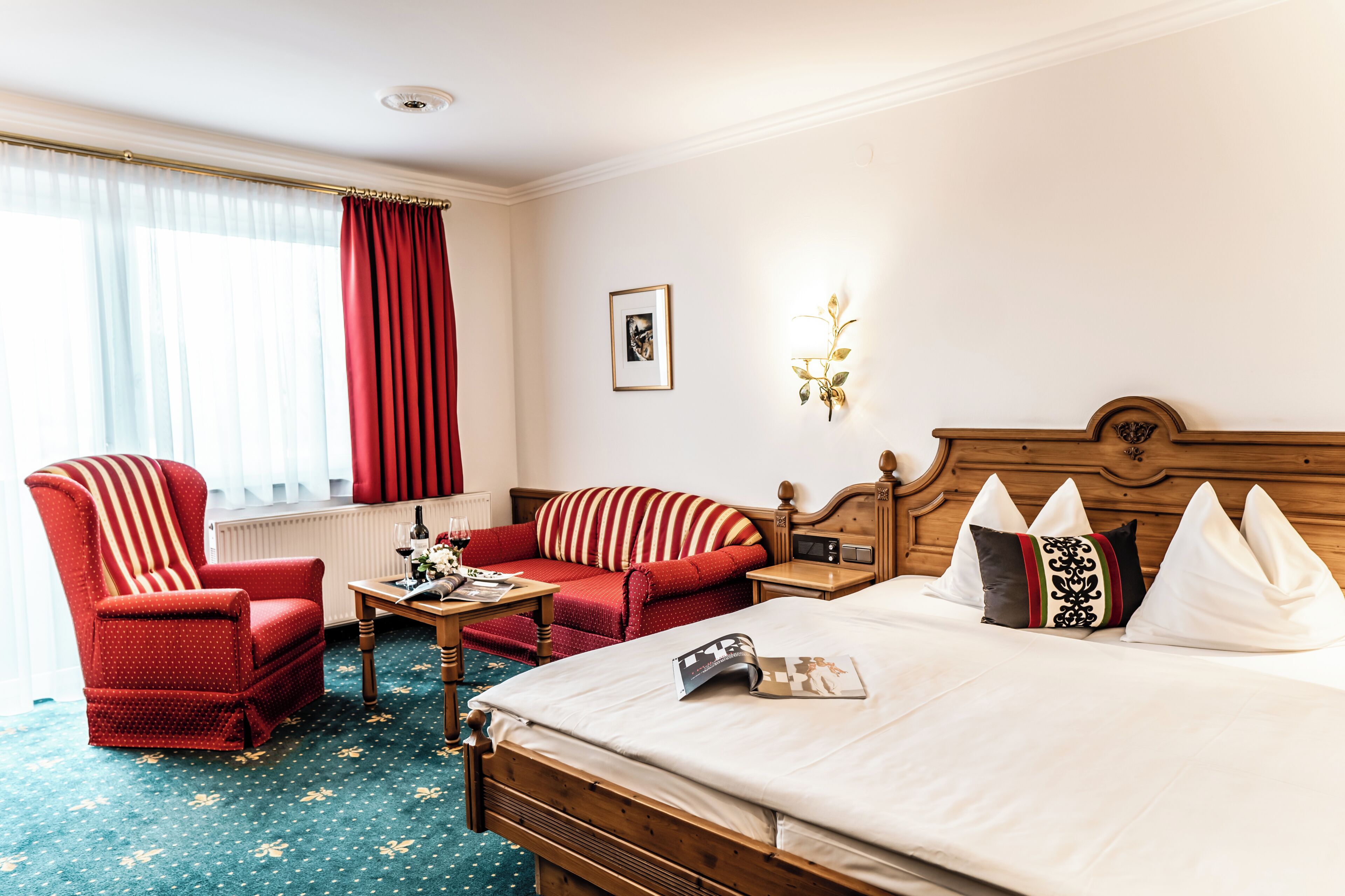 A hotel room with a bed, a red armchair, a sofa, a small table with a wine bottle, and a picture on the wall.