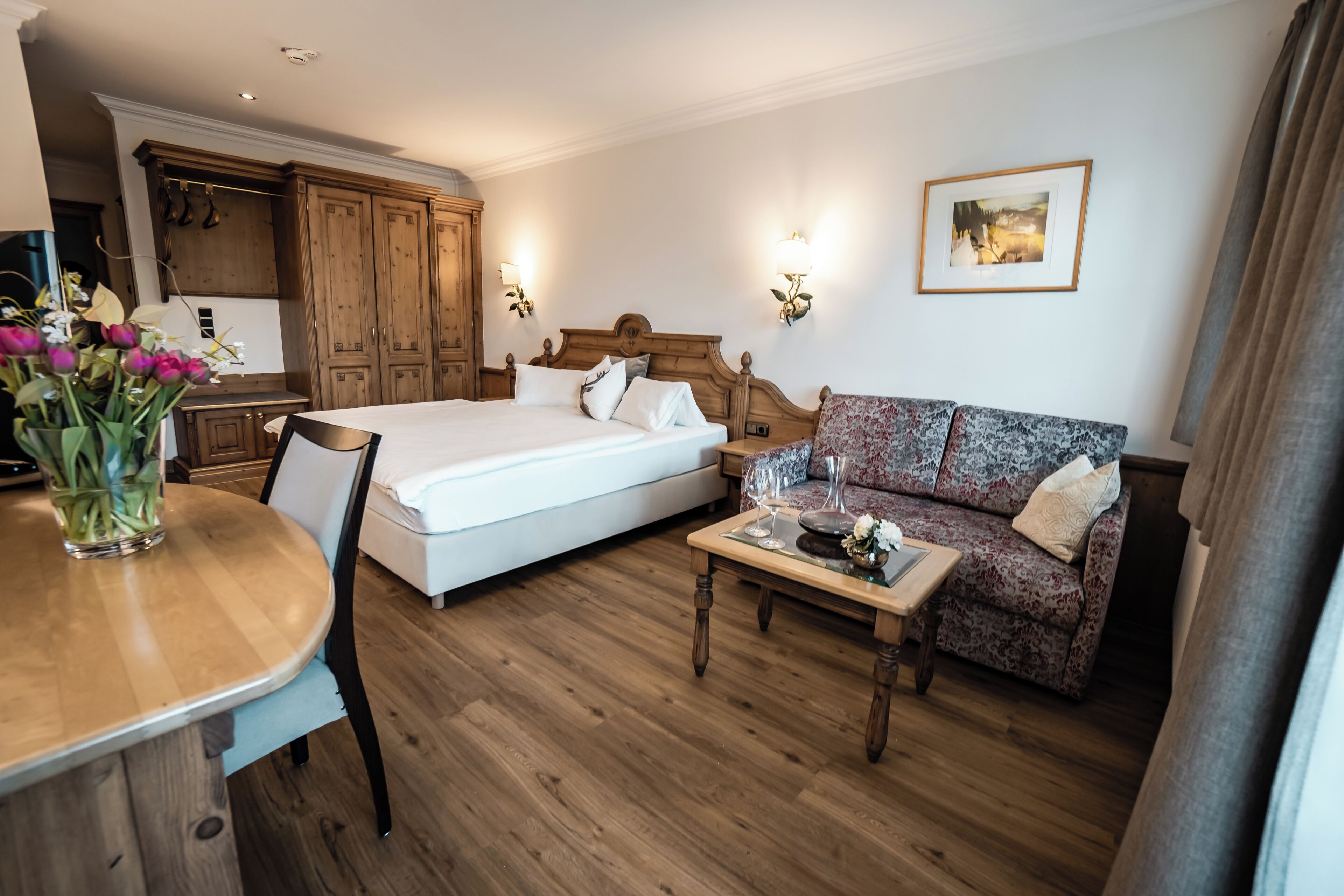 A hotel room with a bed, a sofa, a wooden coffee table, a dining table with a vase of flowers, and a kitchen area.