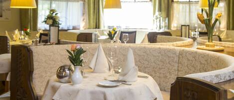 A dining area with tables set for a meal, featuring a patterned tablecloth, a vase with flowers, and a lamp.
