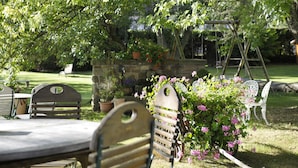 Un área de estar al aire libre con sillas de madera y una mesa, rodeada de abundante follaje y plantas florecientes.