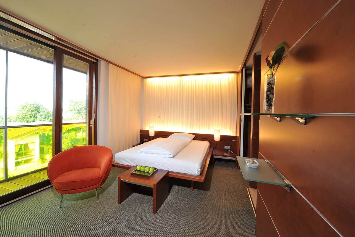 Ein Hotelzimmer mit einem Bett, einem roten Sessel, einem kleinen Tisch mit Obst, einem Nachttisch und Blick auf Grünanlagen durch große Fenster.