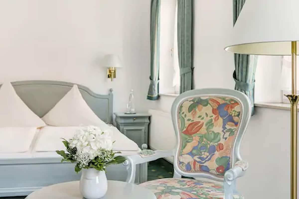 Une chambre d’hôtel comprenant un lit, une chaise recouverte d’un tissu à motifs floraux, une table de chevet avec une lampe et un vase rempli de fleurs.
