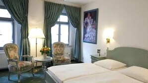 A hotel room with a bed, two chairs, a small table with a lamp, and a painting on the wall.