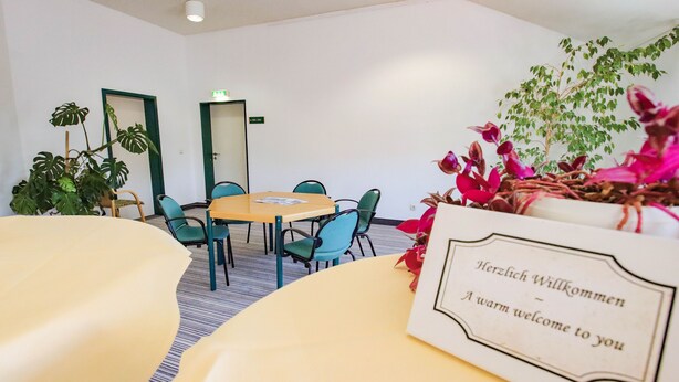 A conference room with a round table, chairs, and a welcome sign. - Hotel Astor in Altenburg (Altenburg)