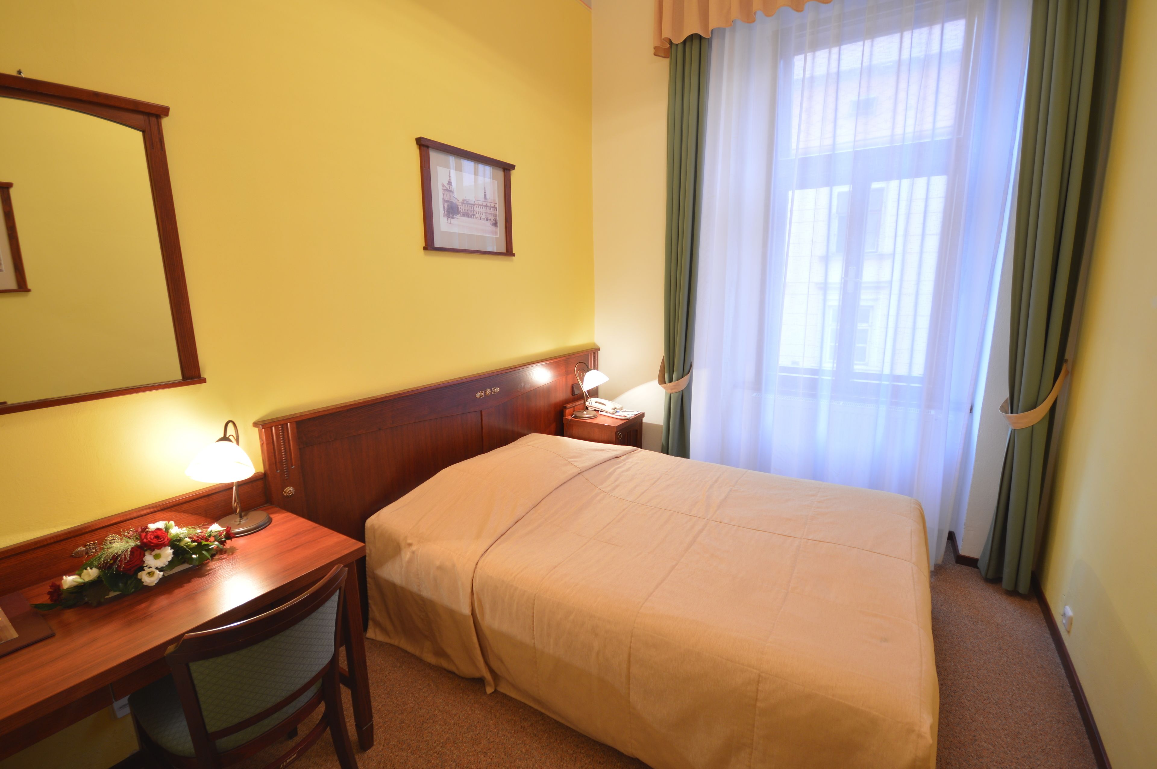 Een hotelkamer met een bed, een bureau met lamp, een stoel, een nachtkastje met lamp, een lijst met afbeelding aan de muur en een raam met gordijnen.