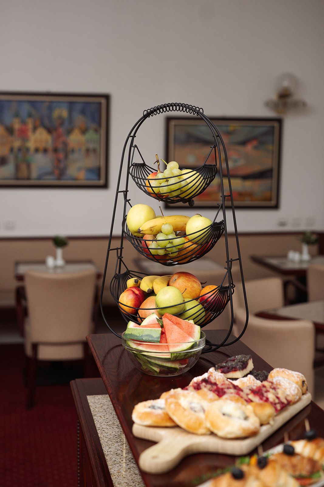 Una frutera de tres niveles sobre una mesa, con una variedad de frutas y una bandeja de pasteles.