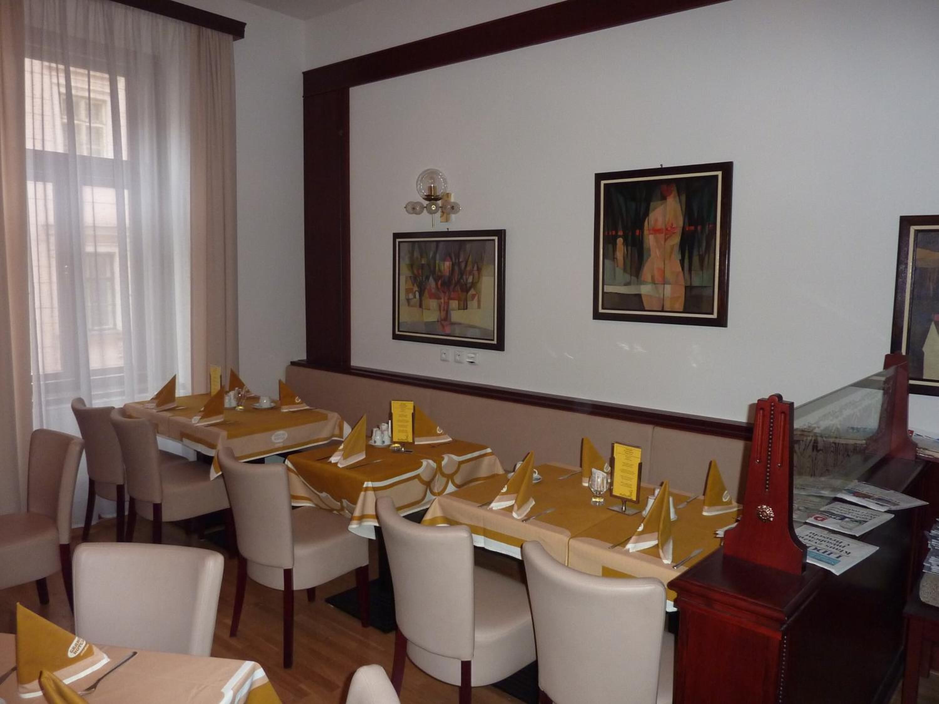 A hotel dining area with tables set for service, framed artwork on the walls, and a window with sheer curtains.