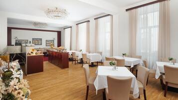A hotel dining area with multiple tables and chairs, a central bar area, and large windows with curtains.