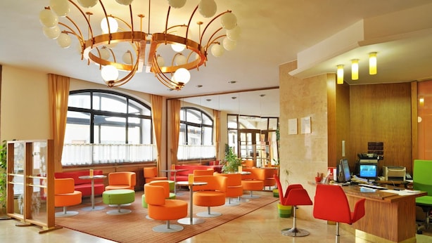 A modern lobby with orange and green seating, a wooden reception desk, and a large chandelier. - Grandhotel Garni (Jihlava)