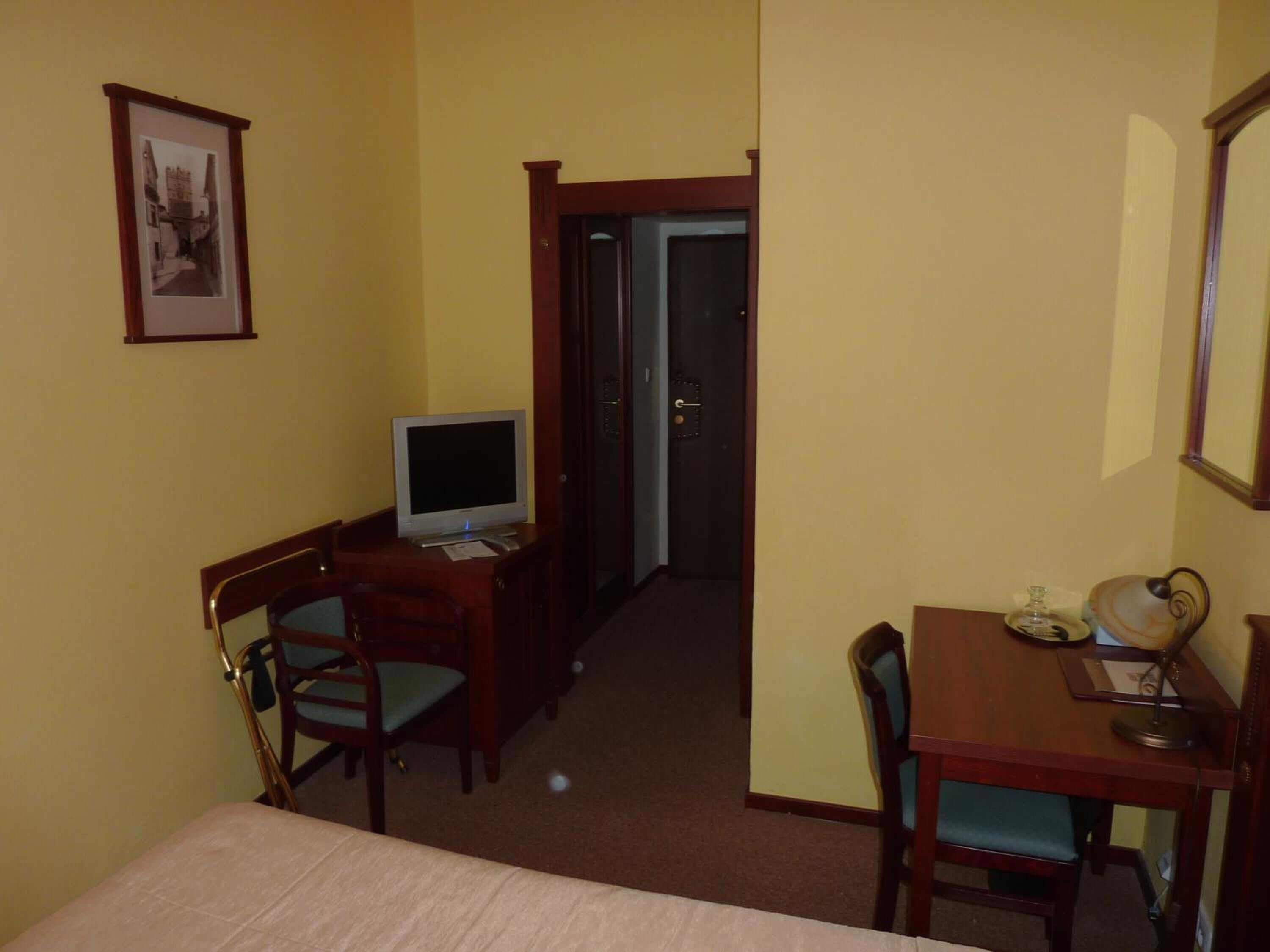 Een hotelkamer met een bed, een bureau met een computer, twee stoelen, een tafeltje met een lamp en een lijstje aan de muur.