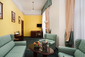 A room with two green patterned sofas, a small table with a fruit bowl and glasses, a desk with a computer, and a television on the wall. - Grandhotel Havel (Jihlava)
