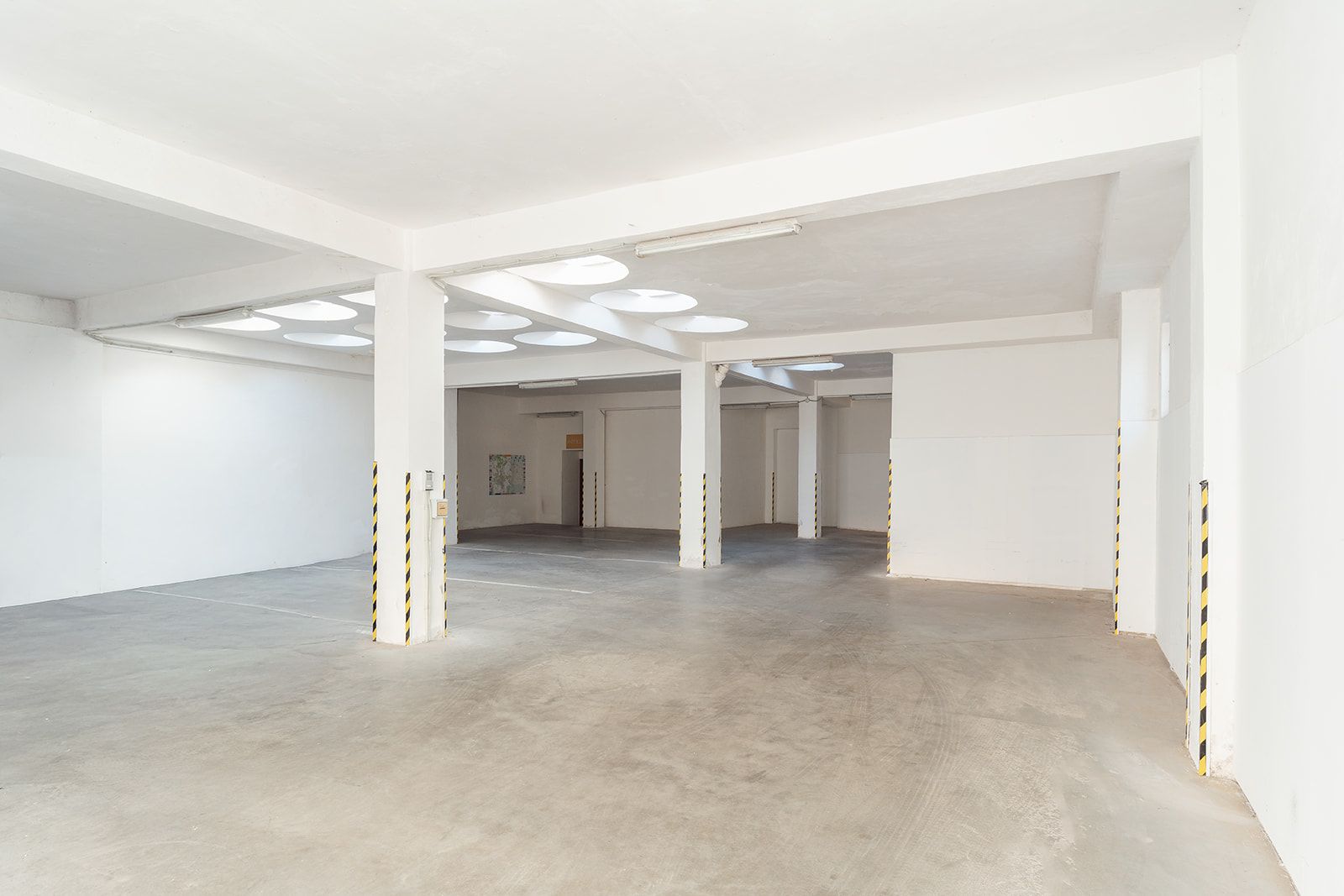 Un interior espacioso y vacío con paredes blancas, pisos de concreto y varias luces en el techo.
