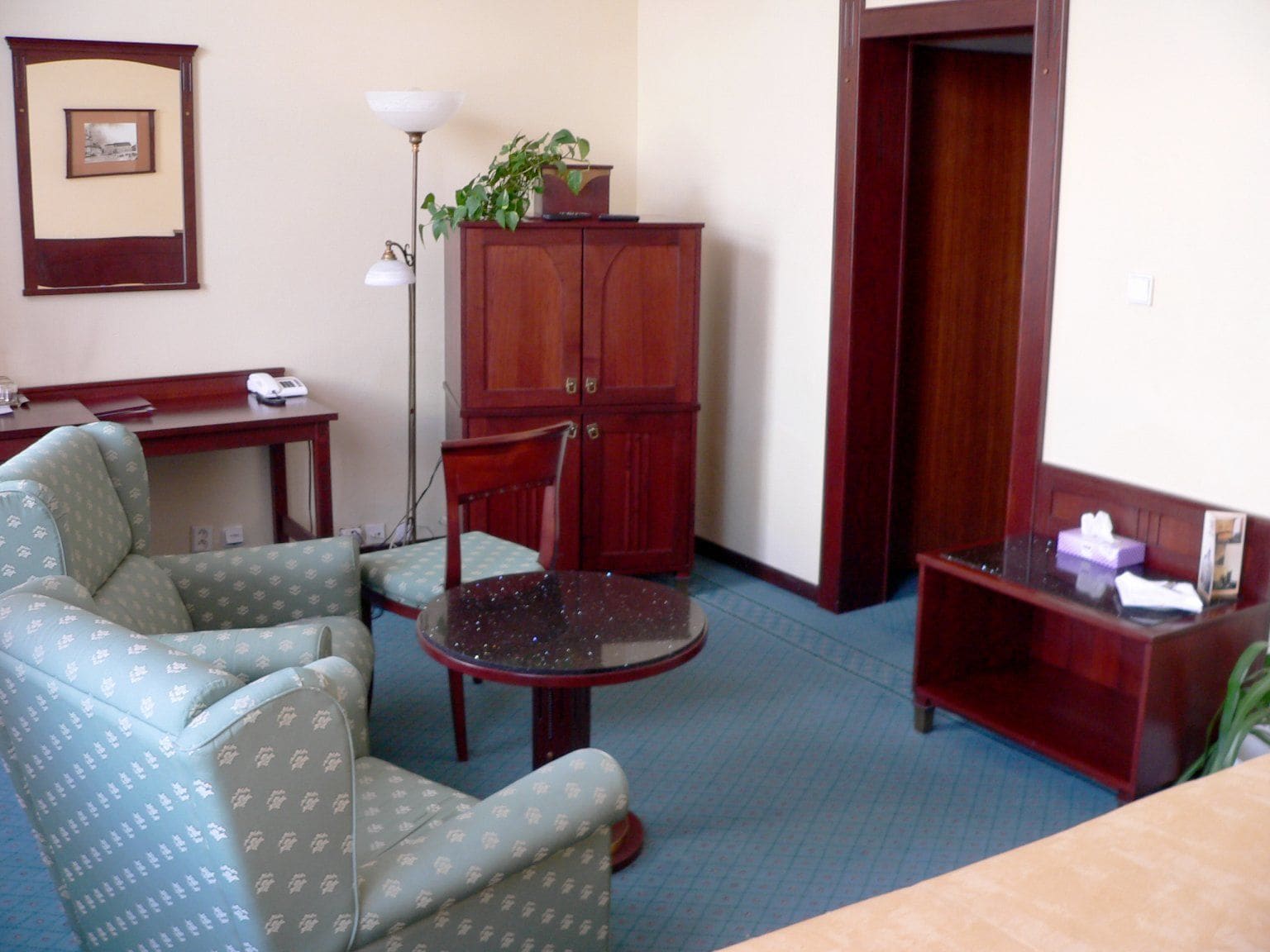 A room with a desk, two patterned armchairs, a small round table, a cabinet, a floor lamp, and a door.