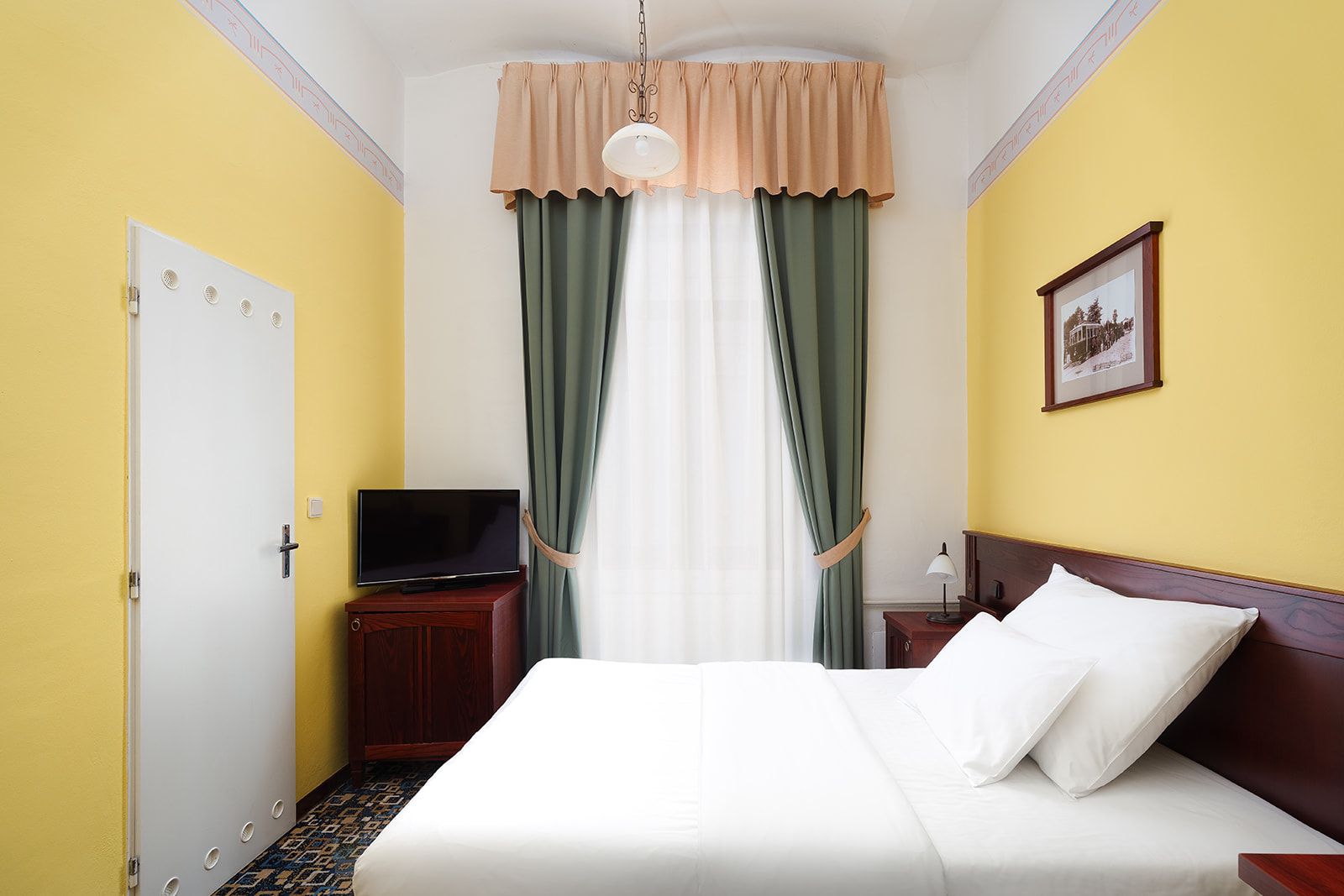Habitación de hotel con una cama, un televisor sobre un mueble de madera, una mesita de noche con una lámpara y un cuadro en la pared.