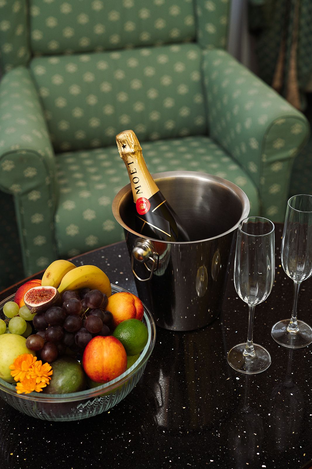 A green upholstered sofa, a glass bowl with fruits and a bottle of champagne, and two empty wine glasses on a black countertop.