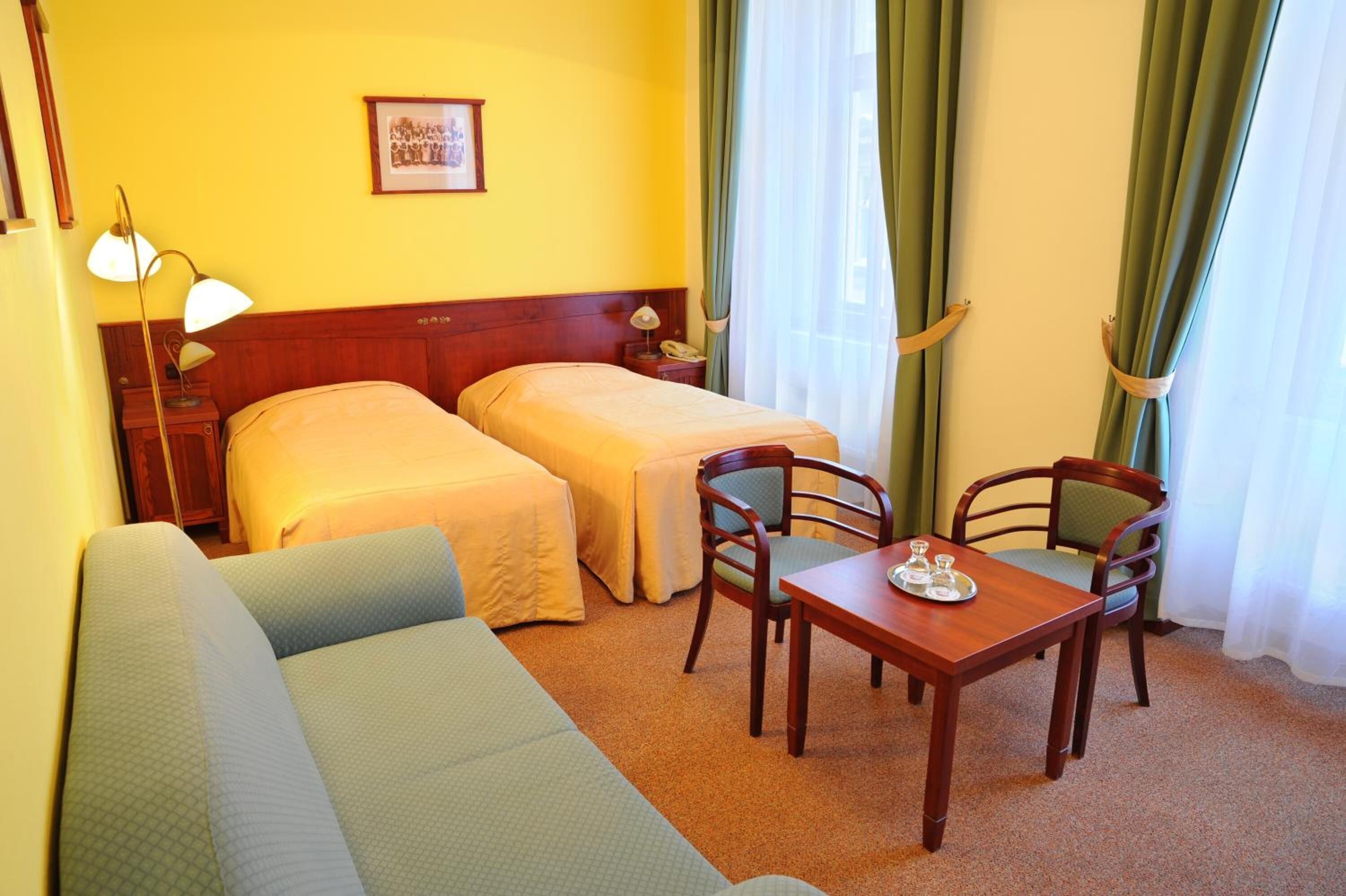 A hotel room with two single beds, a sofa, a wooden table with a tray, and a nightstand with a lamp.