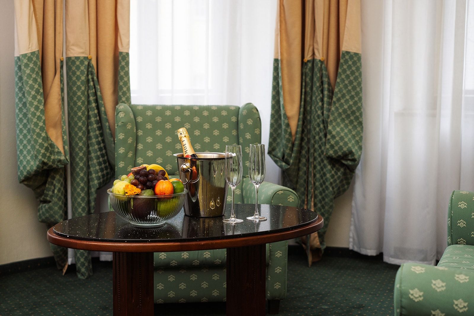 A room with a round table set with a fruit bowl, champagne bottle, and glasses, and green patterned furniture.