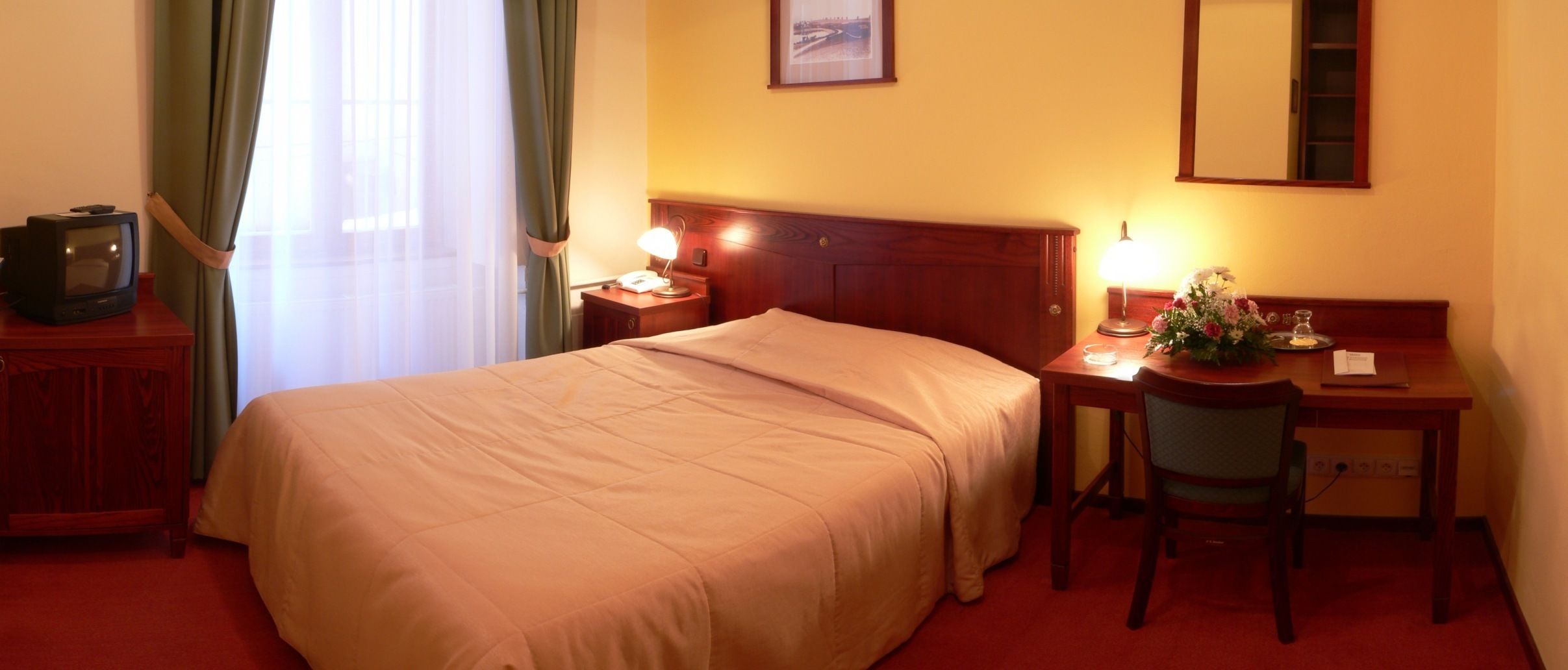 A hotel room with a bed, a desk with a chair, a television, a lamp, and a mirror.