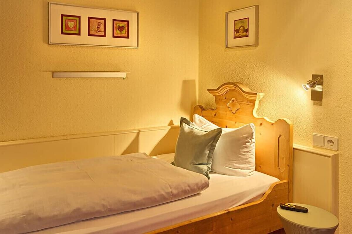 A bed with a wooden headboard, two pillows, a bedside table with a remote control, and framed pictures on the wall.